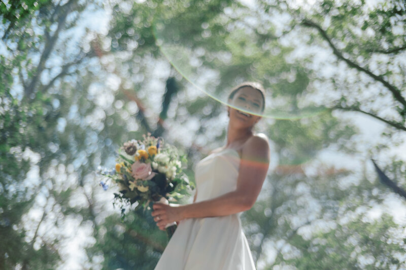 Bride smiles under sunlight, surrounded by trees with a lens flare
