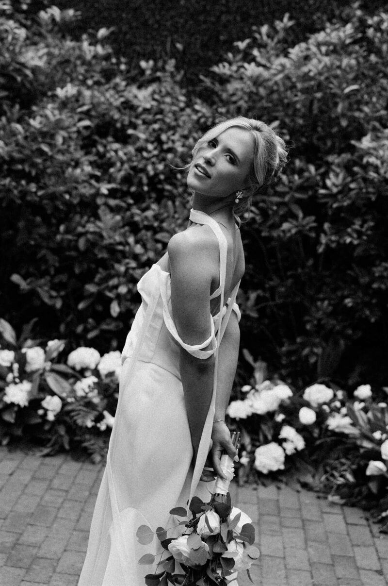 A bride strolls in a flower garden with hands behind her back while looking over her shoulder captured by a film wedding photographer