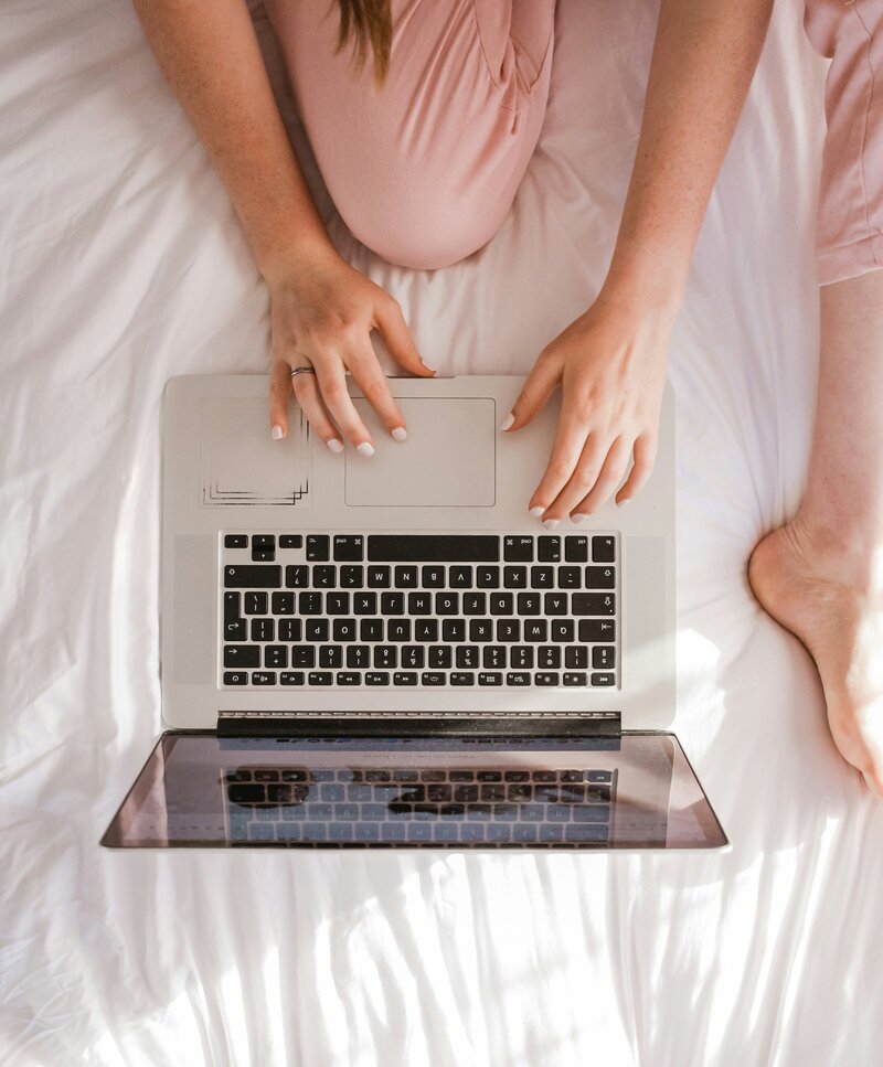 Photo from above of hands typing on a laptop sitting on a white bed