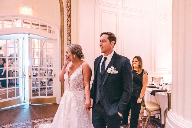 Bride and groom walking excitedly through a ballroom with a smiling wedding planner looking on in the background.