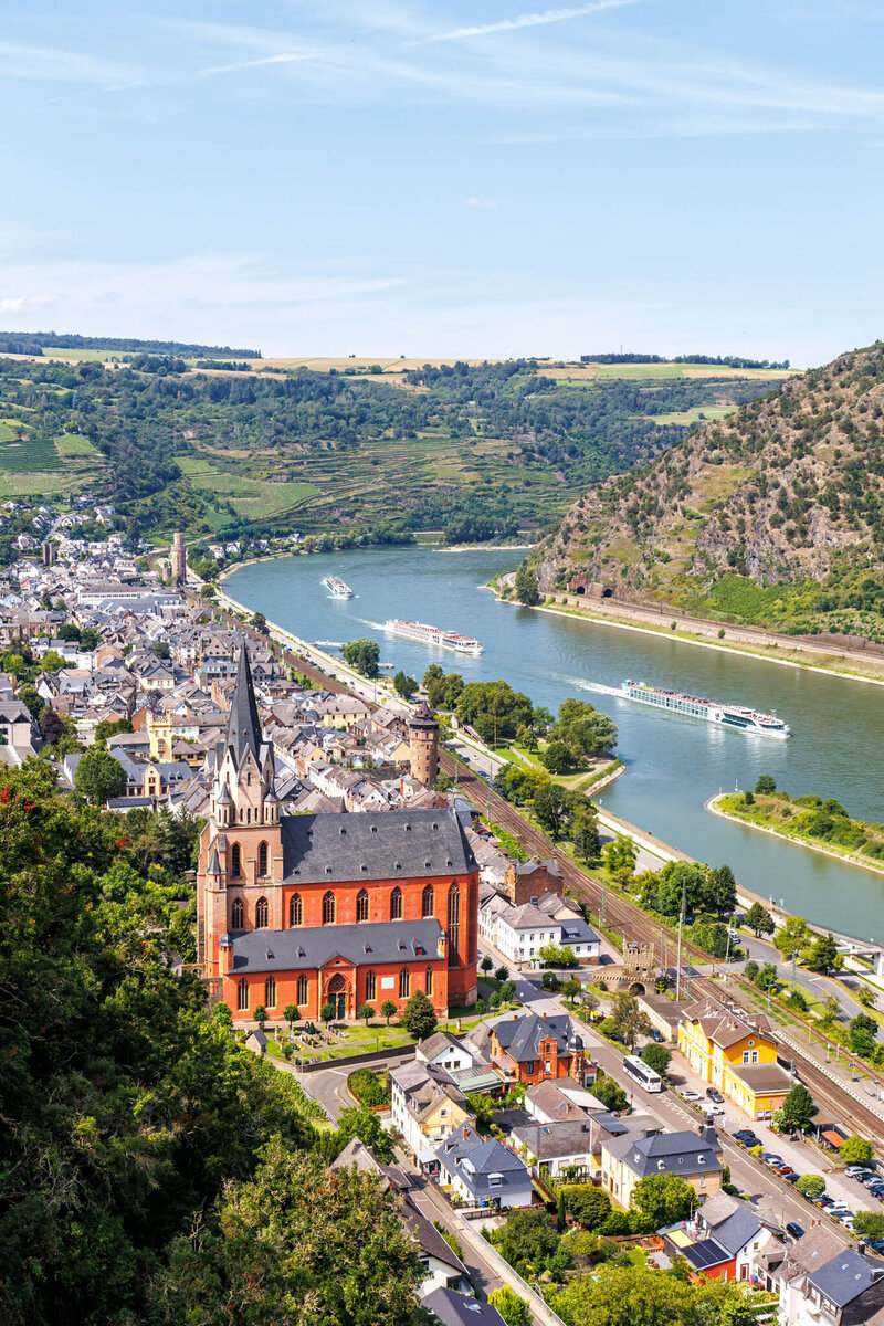 An elevated view of a riverside town featuring a tall red-brick church, clustered houses, and boats cruising along a curving river between green hills.