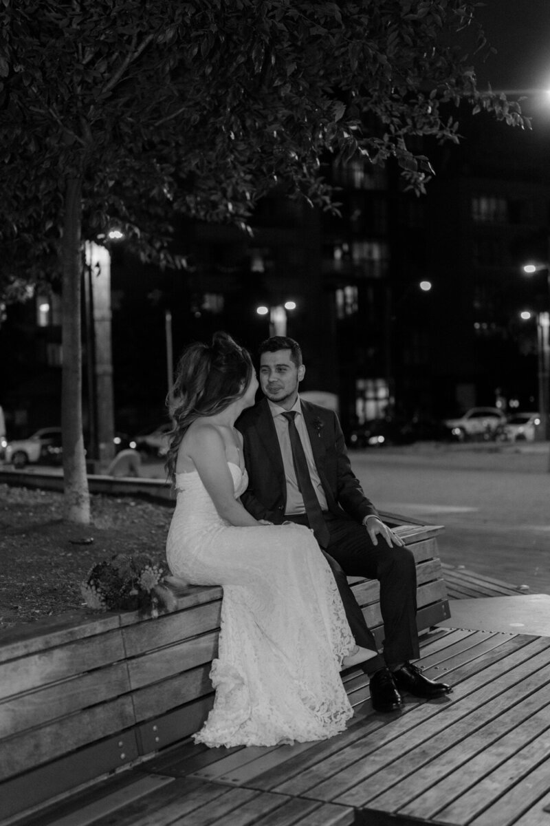 A couple dressed in formal attire sharing a moment on a bench at night.