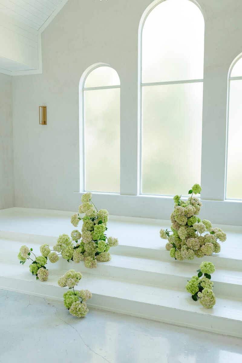A joyful bride in a white satin gown laughs as the groom adjusts her shoe on a marble staircase, a moment of light-heartedness on their wedding day.
