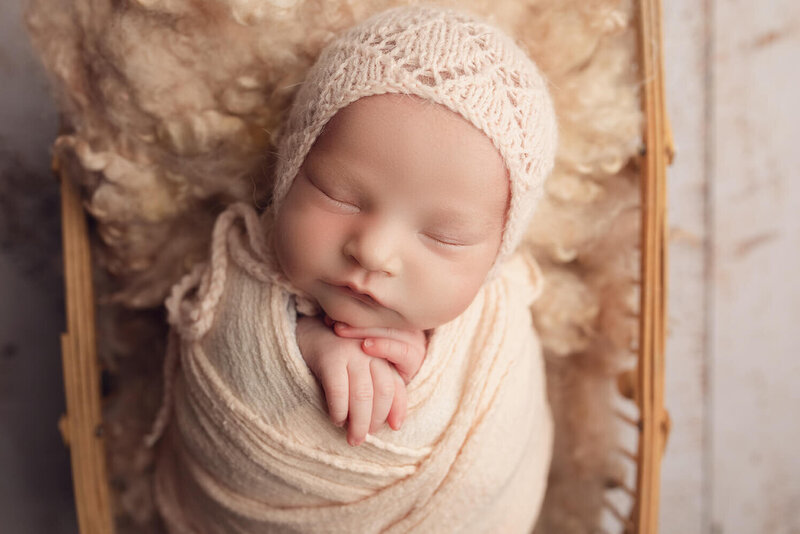 A newborn wrapped in pink wearing a matching bonnet laying in a basket in a studio setting