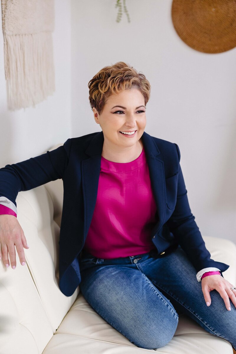 Photographer Teresa Johnson sits on a couch in a white room, wearing a navy blazer and fuschia top.