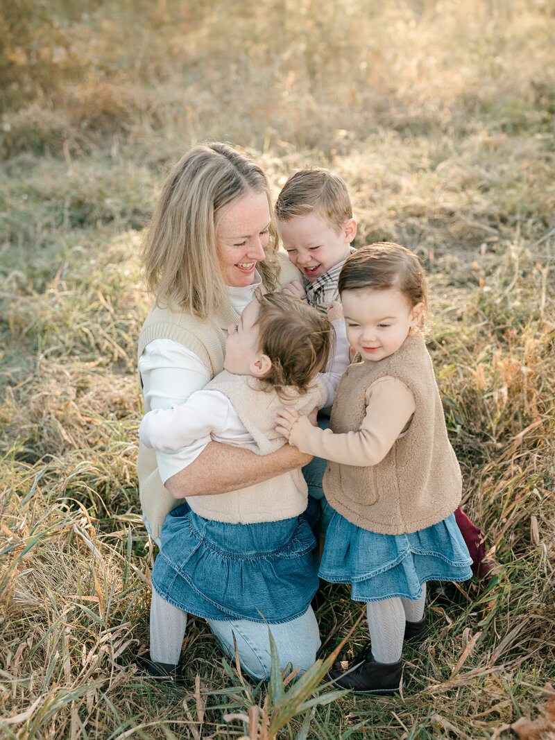 A candid moment of a mother sharing laughter and love with her three young children in a golden field. Dressed in cozy neutrals and denim, their connection shines through in this heartfelt, natural family portrait by Katie Crane Photography.