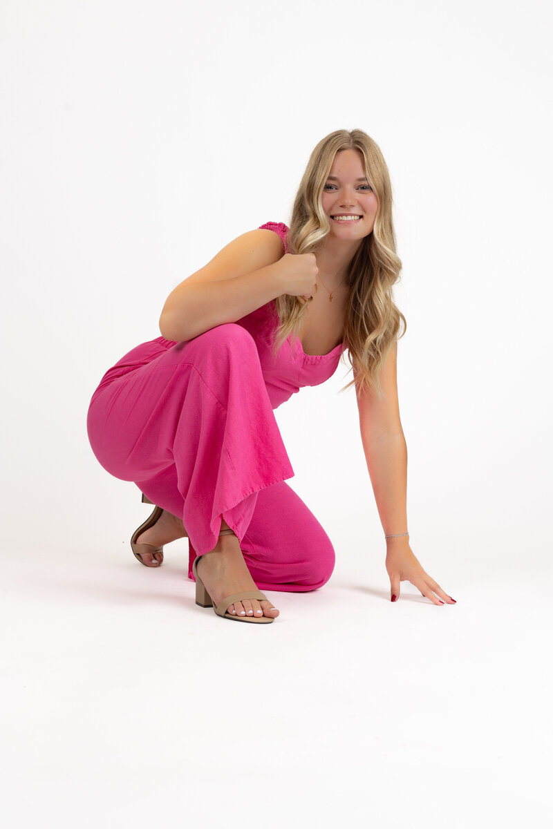 A high school senior girl poses confidently in a vibrant pink jumpsuit against a clean white studio background. With a bright smile and playful energy, this modern senior portrait highlights her personality and stylish fashion choice.