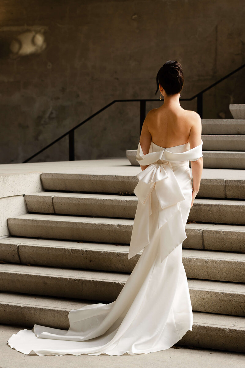 A dramatic photo showing the bow and back details of a bride's gown as she stands on the stairs of the Chateau Laurier wedding venue.  Captured by Ottawa wedding photographer JEMMAN Photography
