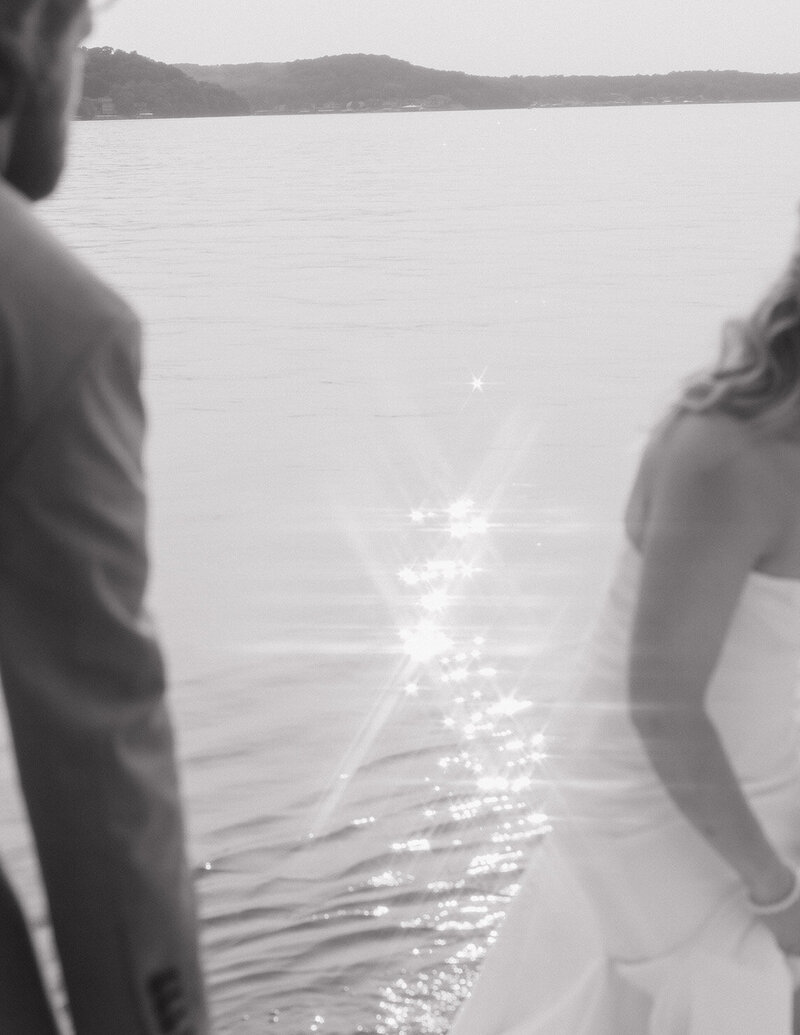 A couple in formal attire stands by a serene lake during sunset. Sunlight sparkles on the water, creating a romantic and tranquil atmosphere.