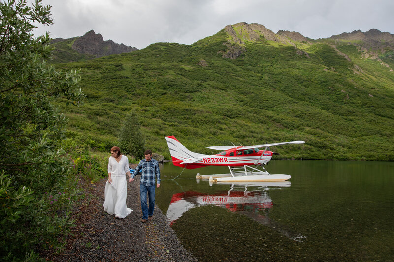 A bride and groom walk along a rocky beach hand in hand as a float plane sits in the lake behind them