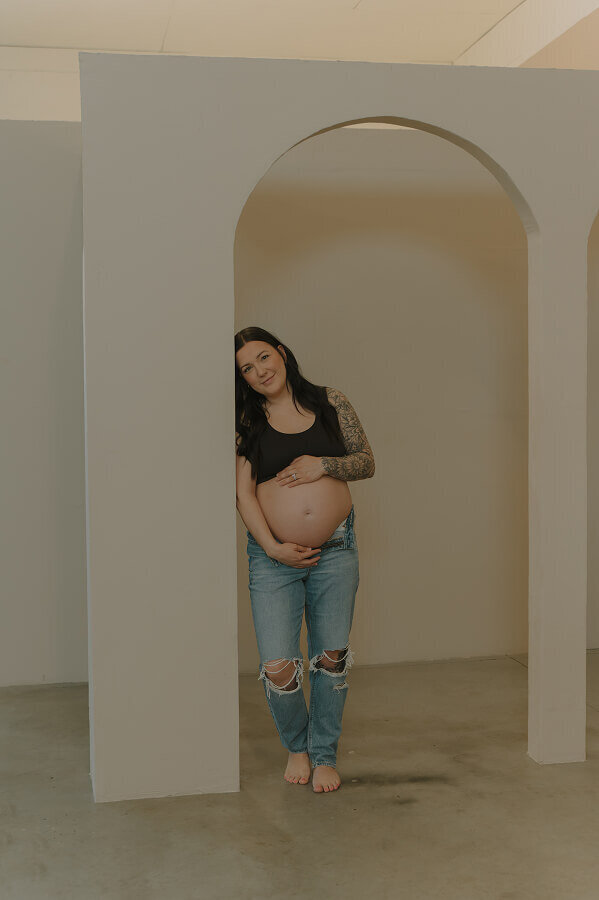 A woman holds her pregnant belly while leaning against a doorway with a rounded arch.