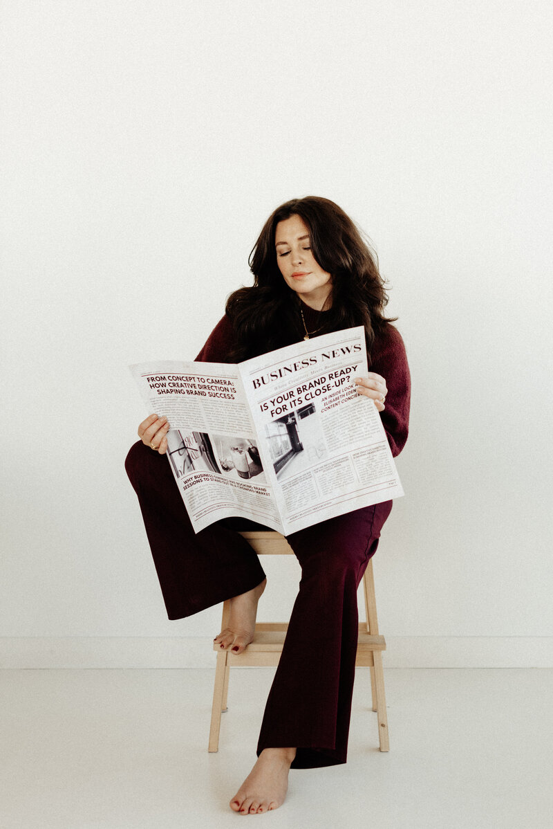 Elisabeth Eden sitting on a stool reading a customized business newspaper branded to her photography agency, blending professionalism with creativity in a personal brand portrait.