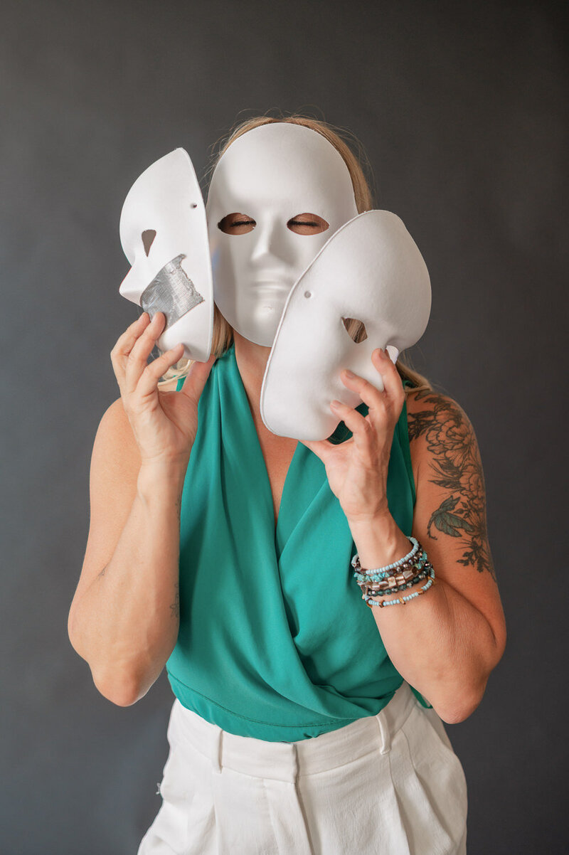 Woman in green top standing in front of black backdrop holding three white masks in front of her face