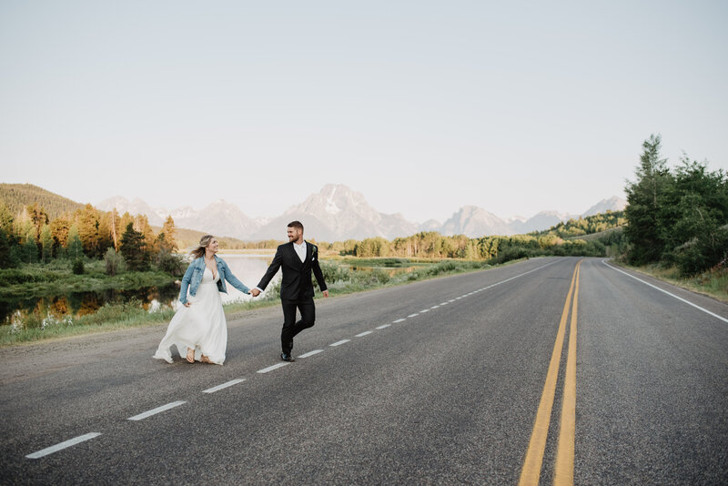 bride and groom holding hands and running across a road in oxbow bend for their jackson hole wedding  as they laugh together for candid wedding pictures