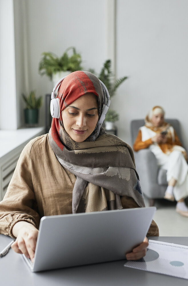 This image shows a woman wearing a red, gray, and brown hijab with headphones overtop, looking down at a laptop in front of her. Several green houseplants are visible in the background of the image.