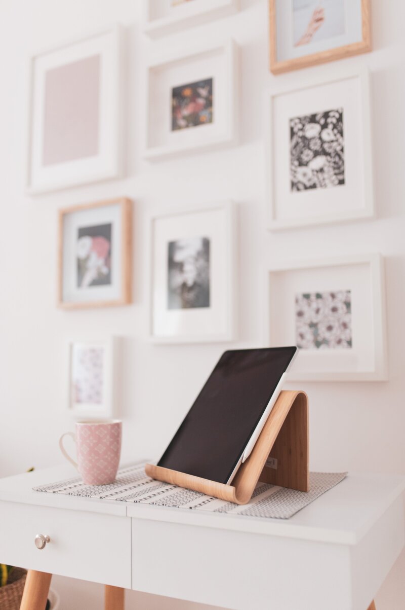 A cozy, stylish workspace with an iPad on a wooden stand, a pink patterned mug, and a soft gallery wall of floral art prints—capturing the calm focus and elevated aesthetic of Design Day Intensives.