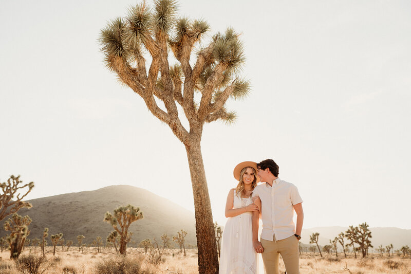 california engaged boho couple  wearing tan and white standin in front of a joshua tree in joshua tree national park. engagement photos in joshua tree national park.