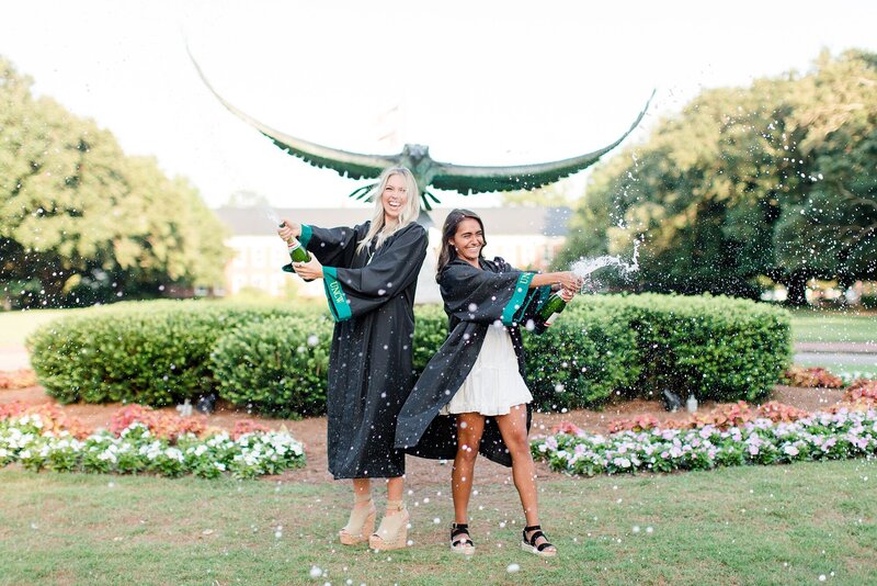 Two women in graduation gowns pop champagne