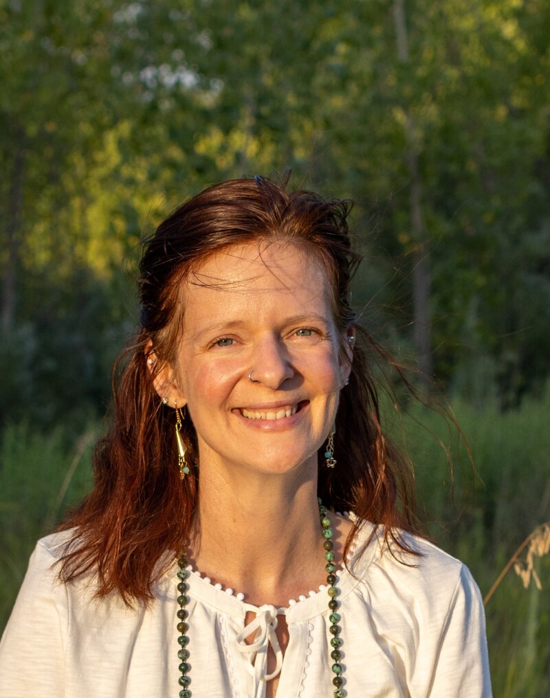 A woman standing in nature in front of a pine tree, smiling and wearing a pink blouse.