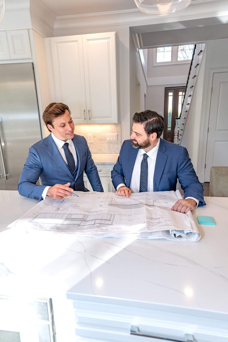 Two men in blue suits sitting at a white marble countertop, discussing with each other while looking at blueprint plans of a  house in Greenwich, CT.