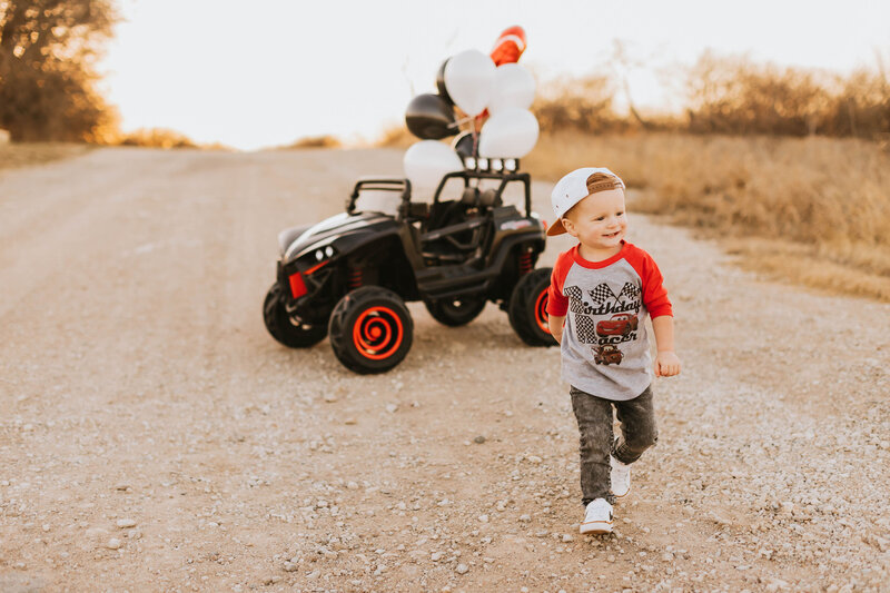 toddler boy walking away from his black jeep that has red, white, and black balloons tied to it