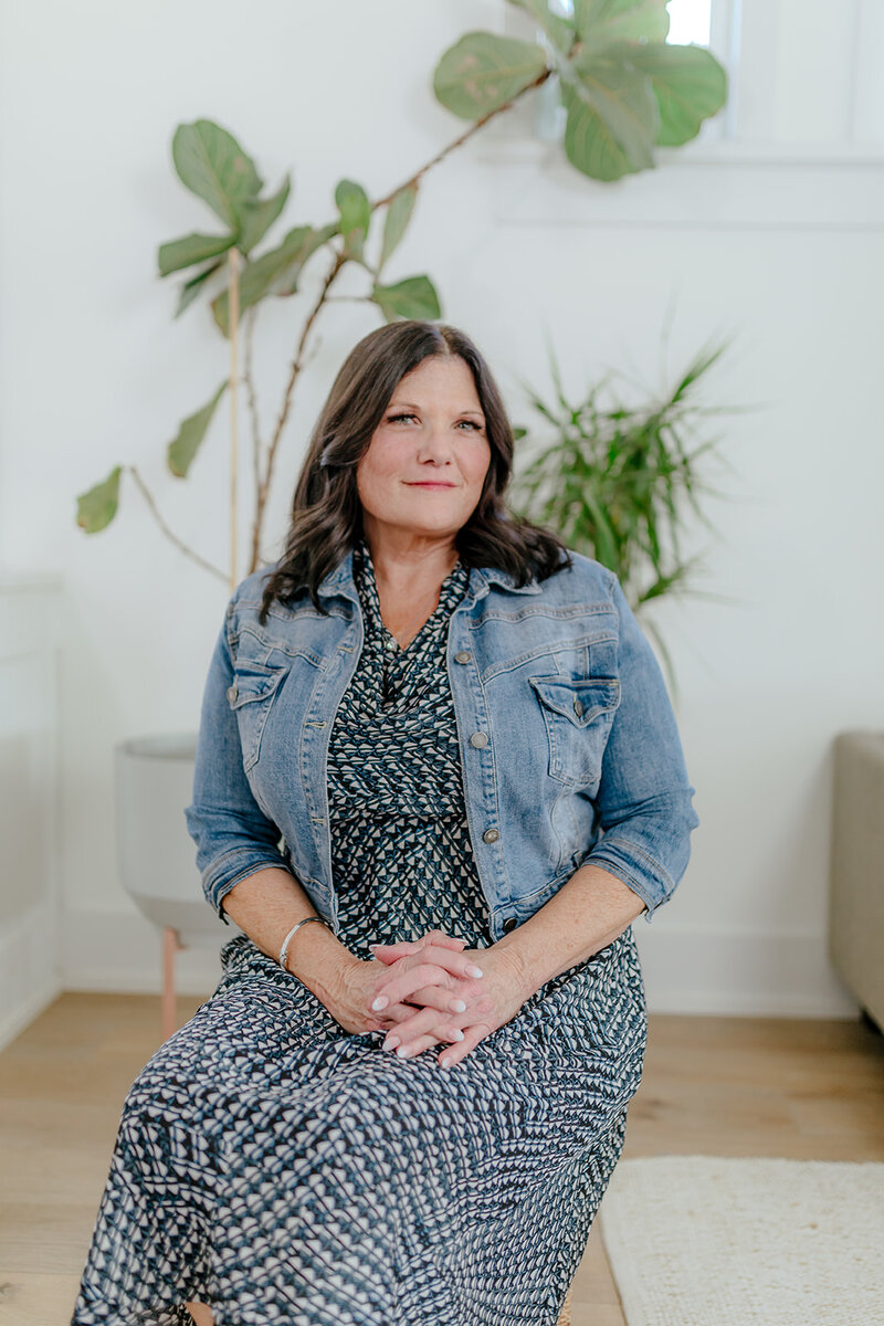 Seated portrait of Susie Schumacher in a patterned dress and denim jacket, hands folded in her lap, surrounded by bright indoor plants in a modern, airy room – Susie Schumacher Life Coach