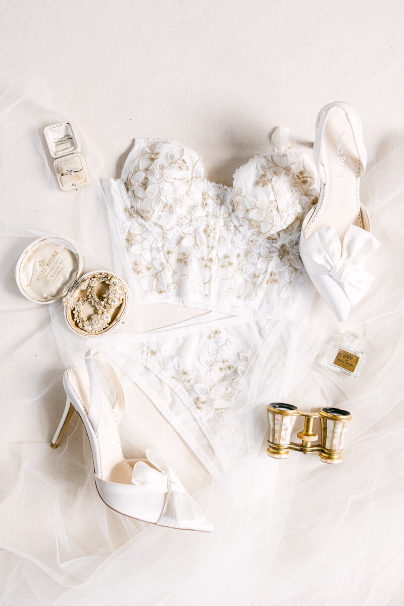 Elegant wedding flatlay featuring a lace bridal corset, white satin heels, heirloom jewelry, and delicate accessories at a luxury Palmetto Bluff wedding.