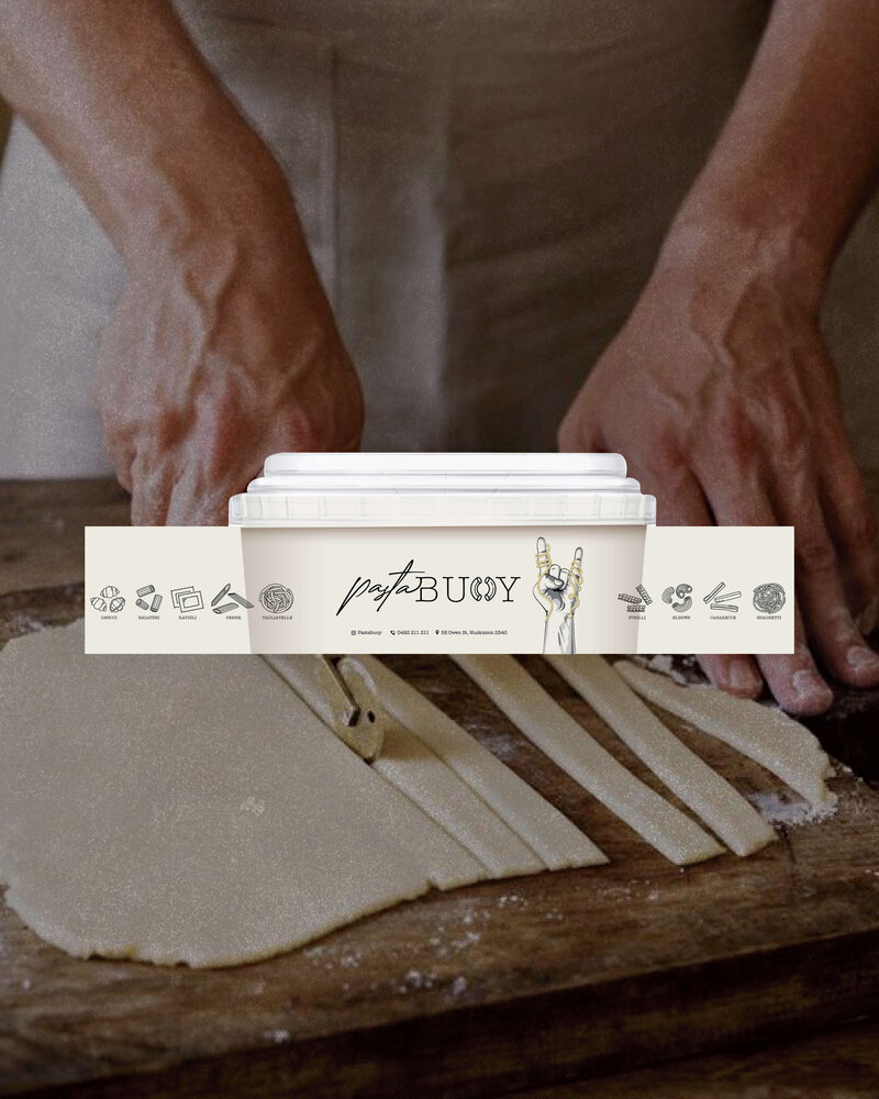 A close-up of hands preparing fresh pasta on a wooden surface, with a Pasta Buoy container placed over the image. The container features playful, hand-drawn illustrations of pasta and utensils, emphasizing the artisan and handmade nature of the product. The rustic scene highlights the traditional craftsmanship involved in making fresh pasta.