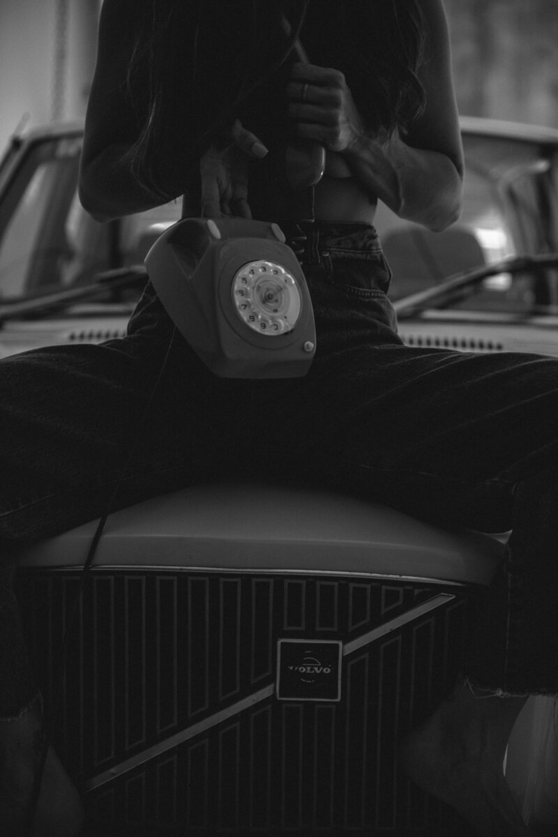 Black and white photo of a woman sitting on a car holding a vintage phone.
