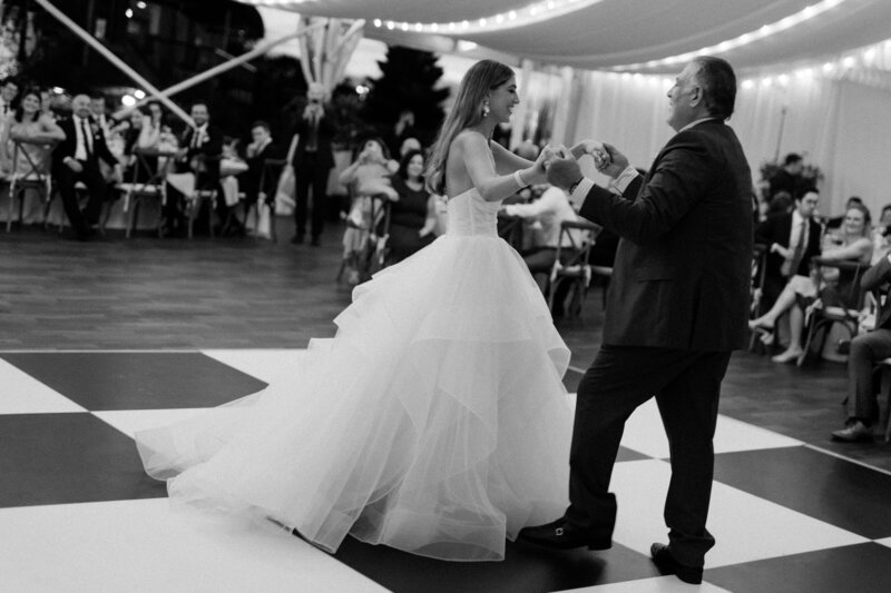 Groom twirls bride beneath wicker lanterns on checkboard dance floor at Glen Foerd Estate in Philadelphia, PA