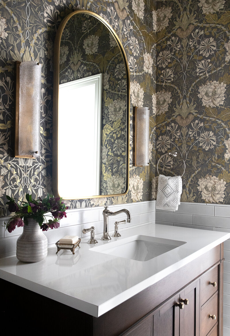 Classic modern transitional powder bathroom vanity with hardwood cabinetry, stone surface countertop and recessed sink. Beautiful floral wallpaper and frosted glass sconce on either side of modern brass mirror