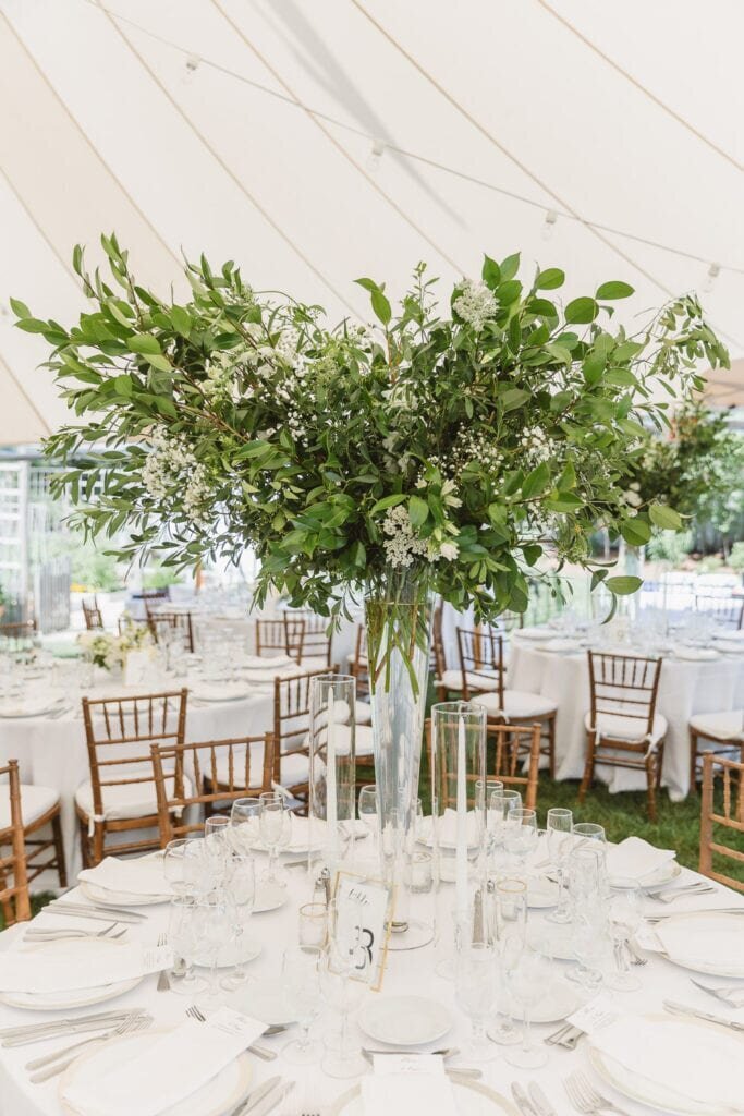 A timeless summer tented wedding celebration in Chester, NJ, showcasing an outdoor reception with lush greenery centerpieces and wooden chairs under a white canopy.