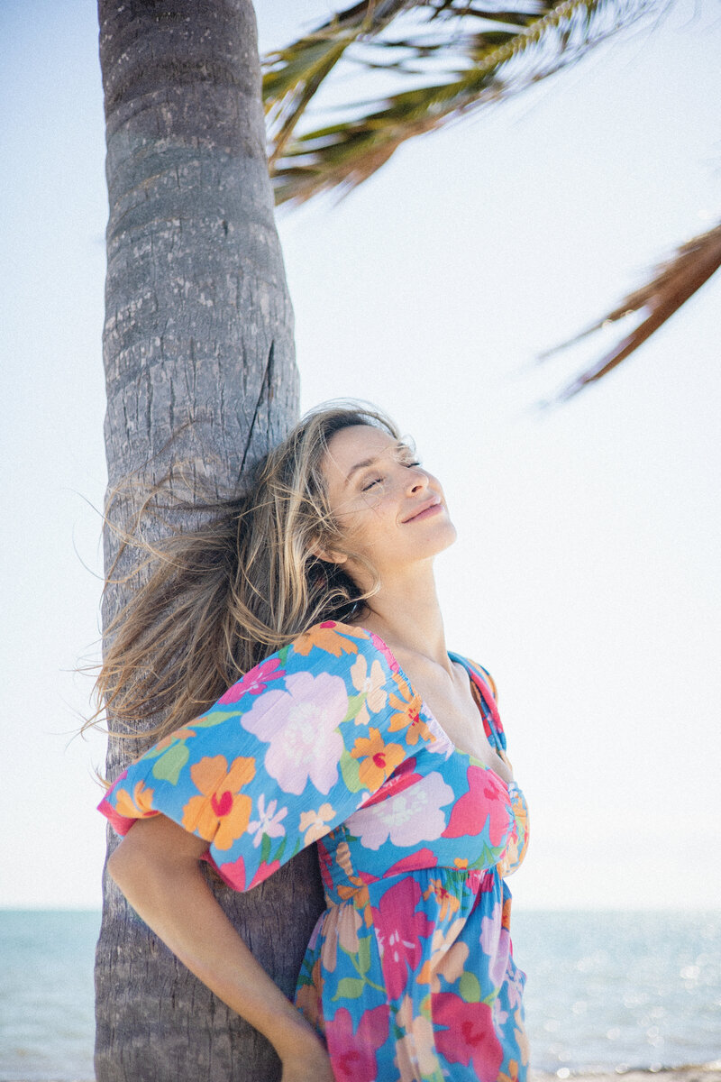 Model wearing swim coverup leans against palm tree