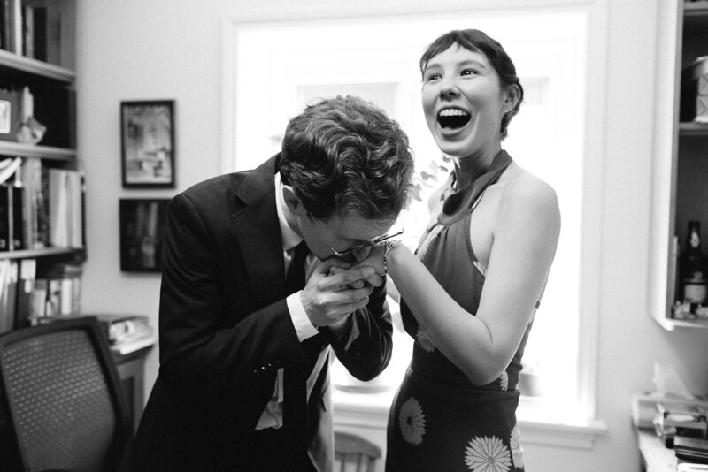 A man in a suit kisses a woman's hand as she laughs in her floral dress. They stand among bookshelves and framed pictures, captured in timeless elegance by a Toronto photographer. Black and white photo.