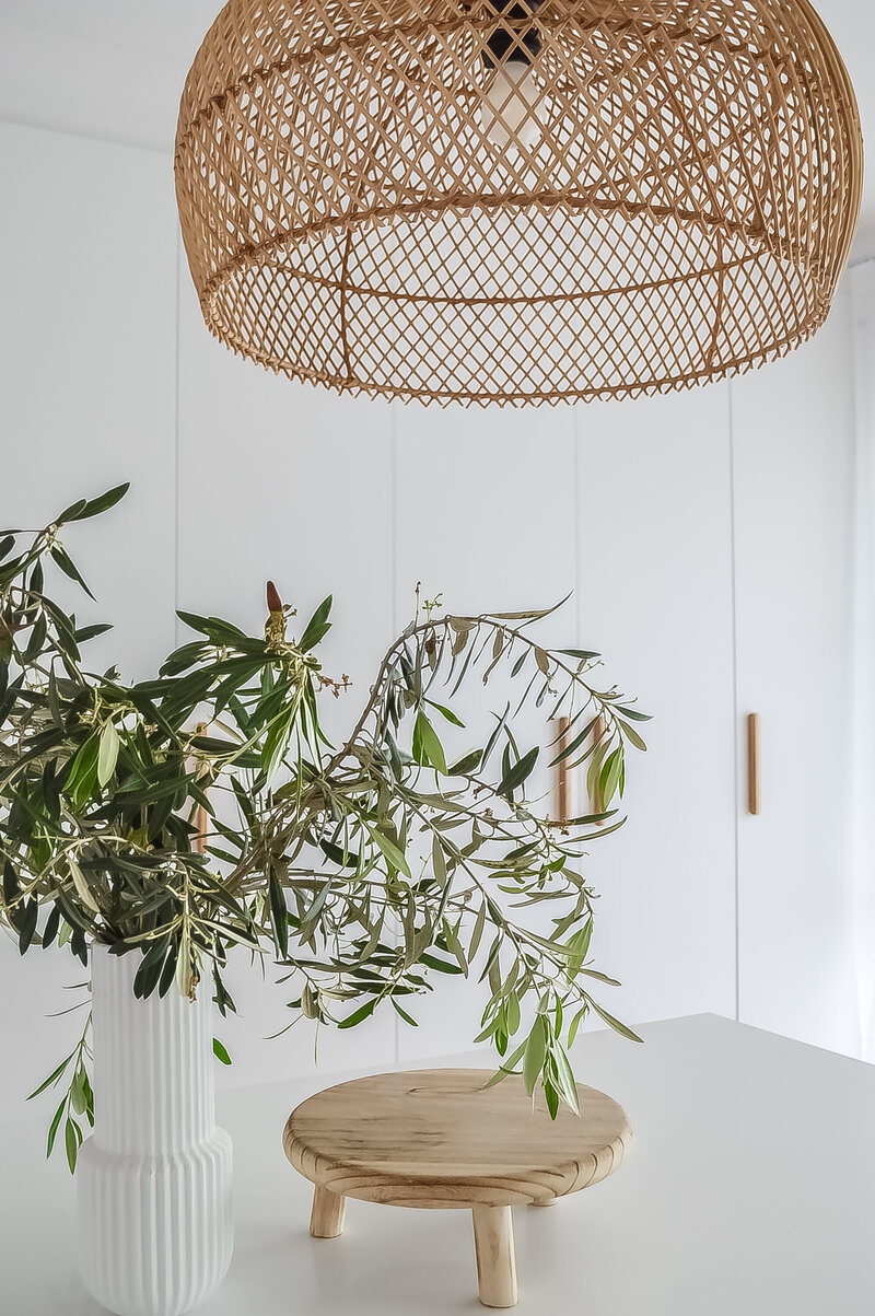 A woven rattan lamp gracefully illuminates the white table, adorned with a plant in a vase and a wooden stand, all nestled within the luxury home design ambiance crafted by a Newcastle interior designer.