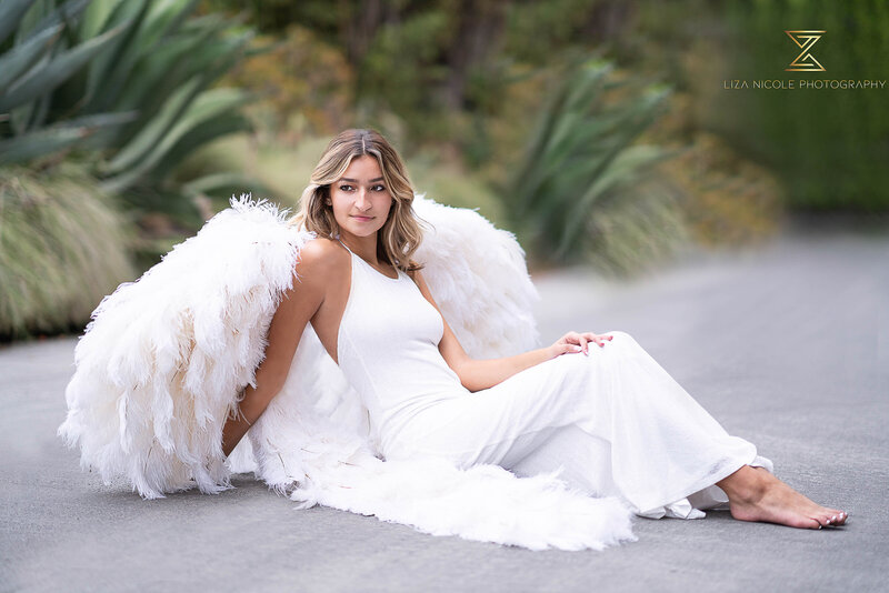 Luxury empowerment photograph captured by top Philadelphia, PA photographer. Woman in long, fitted white dress and full-size feather angel wings is sitting on the ground in a tropical park. She is looking over her shoulder and off in the distance.