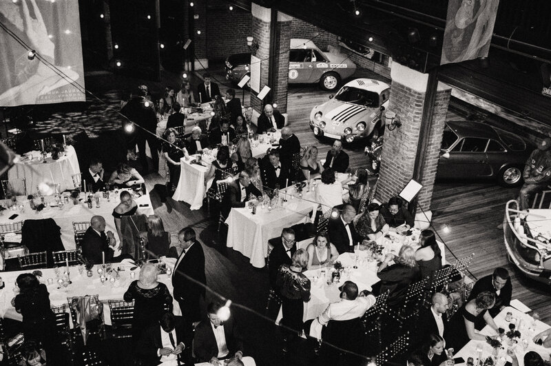 Black and white film photo of a fall wedding reception at the Larz Anderson Auto Museum, showing guests dining beneath string lights with vintage cars in the background — documented by a film wedding photographer.