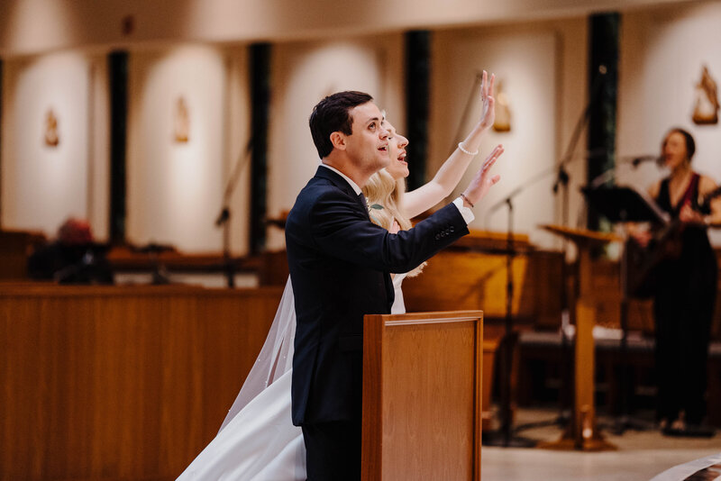 Bride and groom lift their hands in worship as they kneel at the altar in a Catholic church wedding, as wedding musician sings in the background. Photo taken by Orlando Wedding Photographer Four Loves Photo and Film.