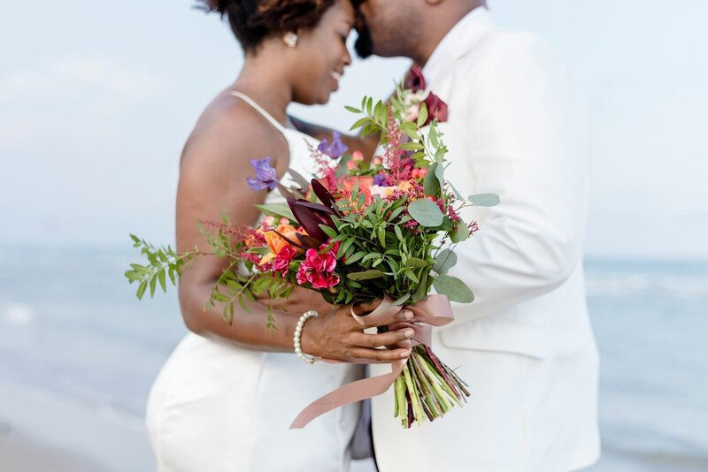 Bride and groom embracing on the beach with colorful bouquet in focus
