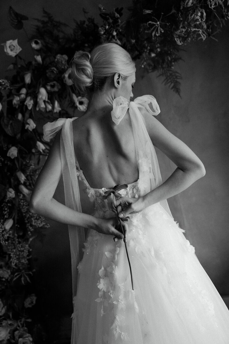 A stunning black and white bridal portrait featuring a bride in a delicate tulle gown, adjusting her dress with a romantic floral backdrop.