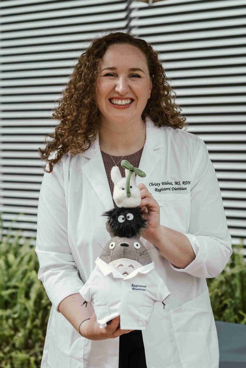 Christy Walowit in a lab coat, smiling while holding a stuffed animal in a bright, cheerful lab setting.