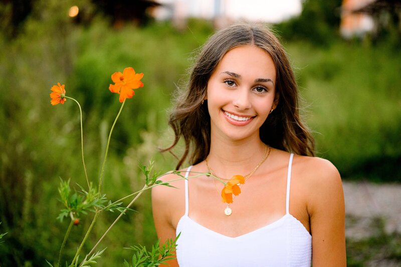 senior stands next to orange flower wearing a white dress