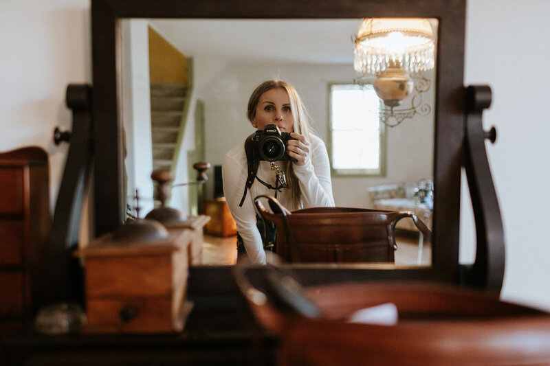 A person captures their reflection in a mirror, camera in hand, surrounded by elegant wooden furniture and a chandelier—Jennifer van Son, photographer.