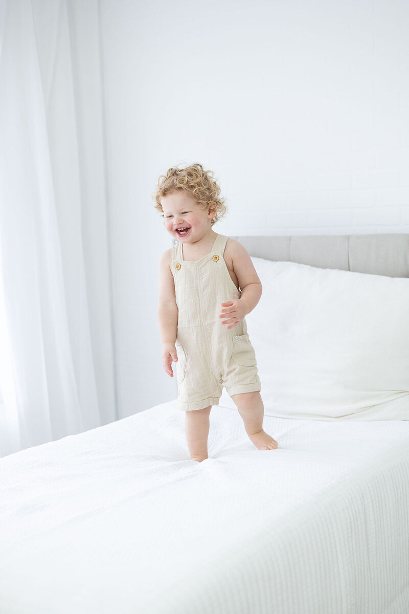 Two year old boy in linen overalls jumping on white bed in studio during NH Baby Photography session with Kathleen Jablonski