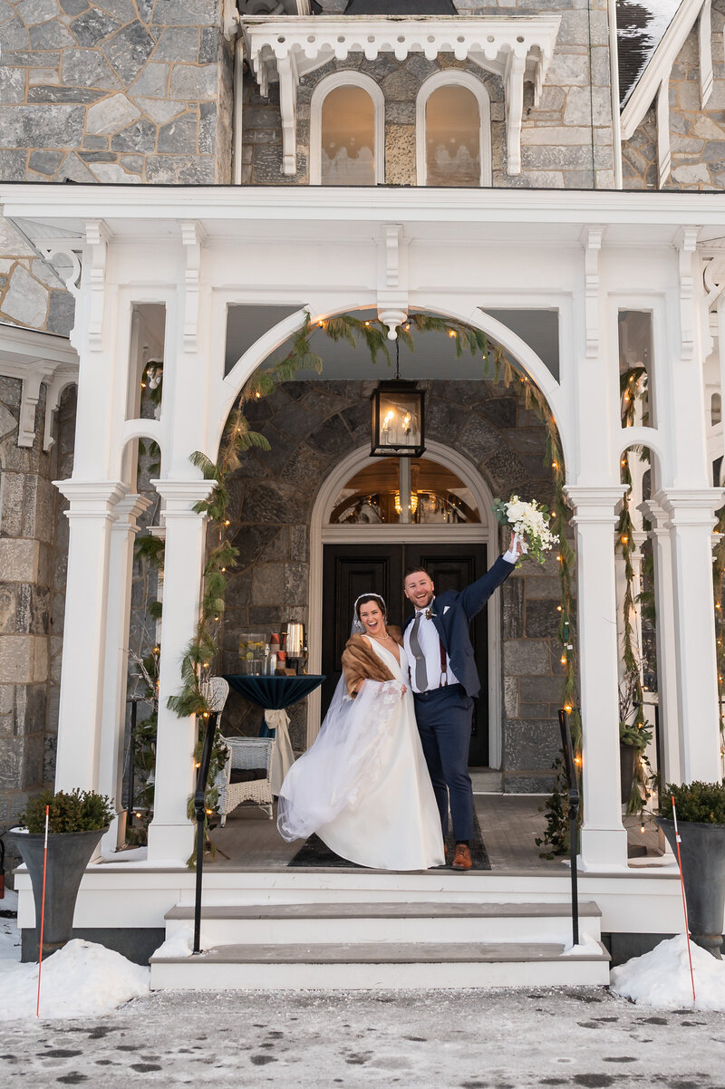 Bride and groom standing on the porch of a large stone mansion with the groom holding the bouquet in the air