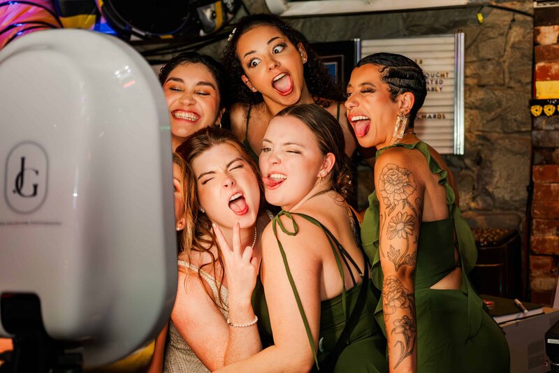 Bride and groom, surrounded by wedding party, taking a photo in front a photo booth, and their faces are illuminated by its light