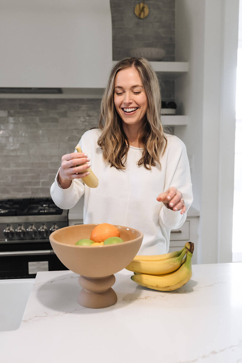 Smiling wellness coach for moms in early motherhood preparing a healthy banana snack in a modern kitchen with a fruit bowl of oranges and limes