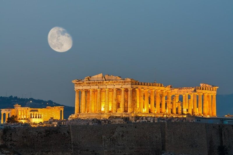 The illuminated Acropolis of Athens stands majestically against a twilight sky, with the glowing full moon adding to its timeless grandeur. This iconic landmark showcases the historical and cultural treasures supported by vacation fundraising for charities.