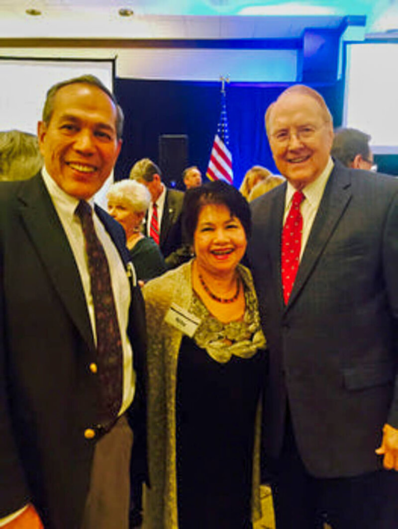 Author and speaker Nita Tin with her husband, Pe Tin, standing alongside Dr. James Dobson of Focus on the Family. Nita wears a black dress with gold leaf embroidery around the neckline and a tan sweater, Pe and Dr. Dobson in dark suits. They are in a large room with many people, with an American flag visible in the background.