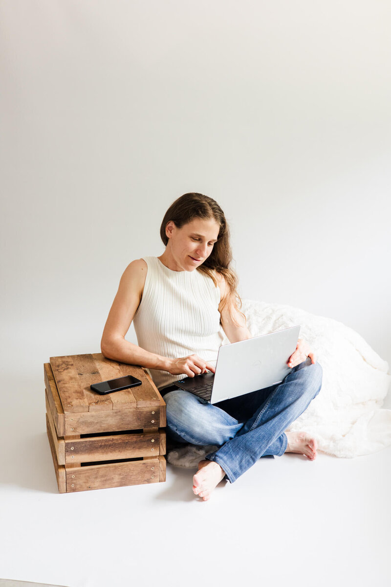 Yeppoon photographer, Julie-Anne, sitting on a photography studio floor dressed in blue jeans and cream top, holding and looking at her laptop.